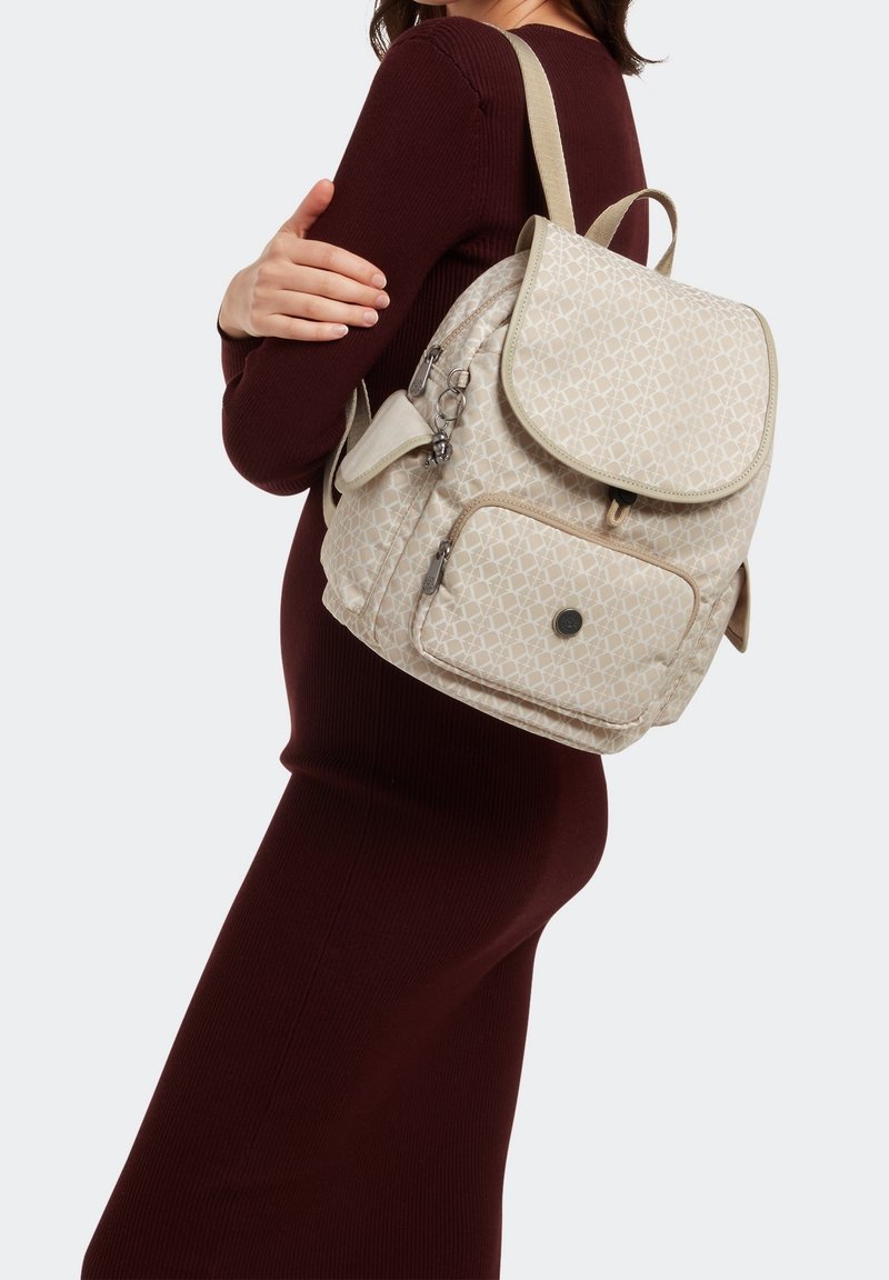 Sac à dos beige à motif avec fermeture à rabat, bretelles réglables, poche avant et ferrures argentées, porté sur une robe bordeaux.