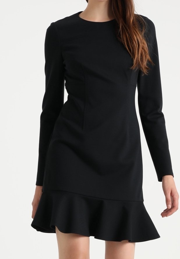 Femme portant une robe noire ajustée à manches longues avec un col rond et un ourlet volanté.