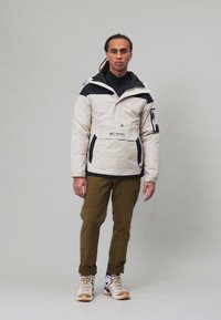 Hvit og svart isolert jakke med glidelås foran, kombinert med olivengrønne bukser. Sneakersene har et flerfarget design.
