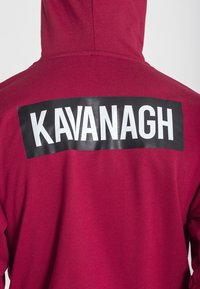 Sudadera color burdeos con un gran parche rectangular negro en la parte posterior que lleva la palabra "KAVANAGH" en letras blancas negritas. Textura suave.