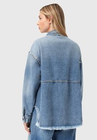 Γυναίκα με μακριά ξανθά μαλλιά, φοράει ένα φαρδύ, ανοιχτό γαλάζιο τζιν μπουφάν με ξεφτισμένα άκρα και διακοσμητικά καρφιά στα πάνω μανίκια, φωτογραφημένη από πίσω.