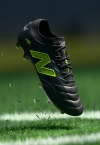 Melnas un neona zaļas New Balance futbola kedas gaisā virs zāles ar izkaisītiem asmeņiem un baltu laukuma līniju zemāk.