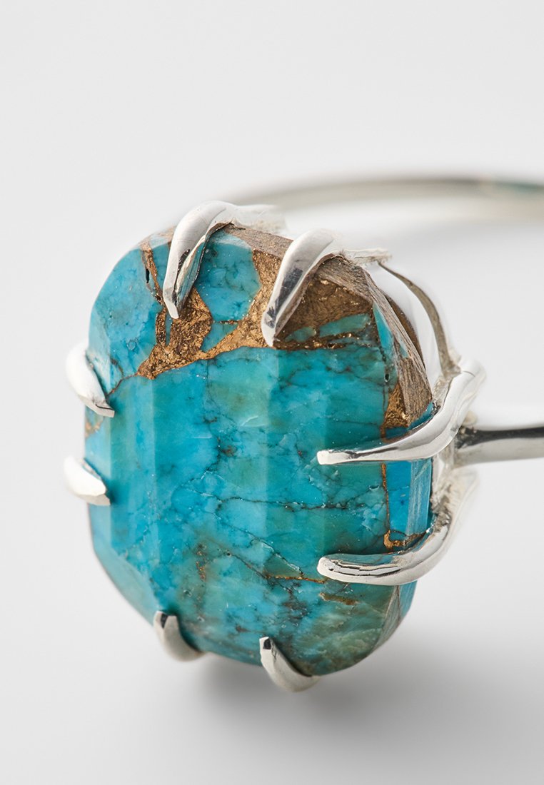 Anillo de turquesa con una piedra rectangular facetada engastada en garras de plata. La piedra exhibe tonos azules con vetas doradas y una superficie pulida.
