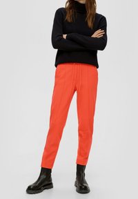 Rote schmal geschnittene Hosen mit Seitennähten, elastischem Bund und schwarzen Stiefeletten. Das stehende Model trägt einen schwarzen Pullover mit langen Ärmeln.