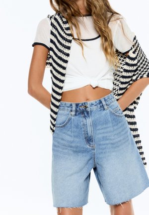Femme portant un t-shirt court blanc, un short en jean clair taille haute, et un pull rayé noir et blanc posé sur les épaules.