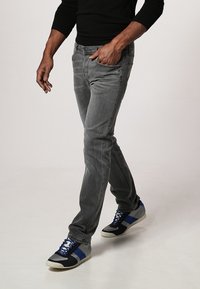 Jeans di denim grigio con vestibilità slim, caratterizzati da una texture sbiadita. Abbinati a sneaker sportive nere e blu. Modello che cammina contro uno sfondo neutro.