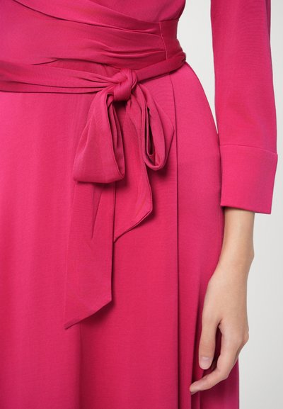 Φούξια φόρεμα με φαρδιά, δεμένη ζώνη στη μέση. Το ύφασμα φαίνεται ομαλό και απαλό, με μακριά μανίκια και ένα ρέον τμήμα φουστίνας.