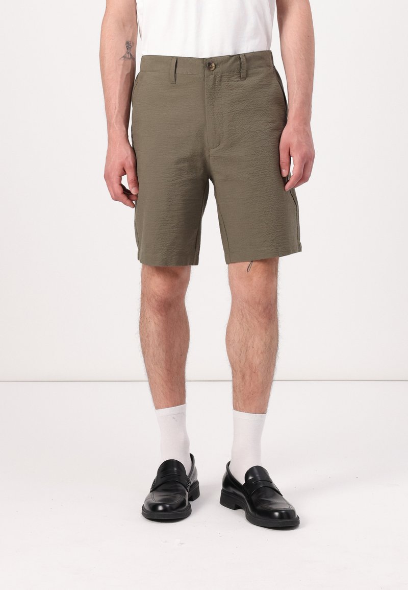 Shorts vert olive avec une texture froissée, fermeture à bouton, deux poches latérales et une coupe droite, associés à des mocassins noirs et des chaussettes blanches.
