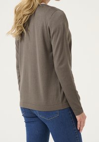 Braune gestrickte Strickjacke mit langen Ärmeln, rundem Ausschnitt und lockerer Passform. Über blauen Jeans getragen, die eine glatte Textur zeigen.