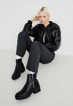 Mustad must saadikavööd, mis on varustatud paksu tallaga, sileda nahapinnaga, külgsepose ja plokknõrklehiga. Kanda hallide pükstega ja valgete sokkidega.