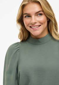 Blouse verte à col montant en tissu texturé, avec des manches bouffantes et un design doux et fluide. Gros plan sur le col et les détails des épaules.