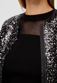 Svart paljetbomberjacka med dragkedja, som har ett texturerat svart nätöverdrag och en åtsittande svart topp under.