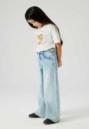 Fiatal lány hosszú sötét hajjal lefelé néz, fehér tigris mintás pólót, világoskék bő szárú farmert és fekete cipőt visel egyszerű háttér előtt.