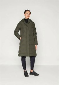 Kvinde iført en knælængde olivengrøn puffjakke med hætte, sorte leggings og sorte sneakers, stående foran en ensfarvet baggrund.