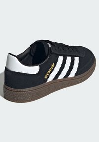 Sapatilha de camurça preta com três riscas brancas, logotipo dourado "SPEZIAL" e sola de borracha castanha com padrão texturizado. Design de atacadores.