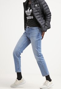 Person som bär en svart Adidas-jacka och tröja, ljusblå jeans, svarta strumpor och vita sneakers, står med ett ben framåt.