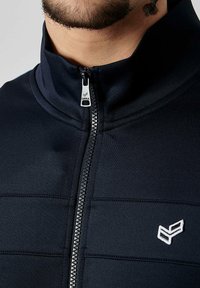 Veste zippée navy avec un col haut, une texture lisse, des coutures horizontales et un petit logo blanc sur la poitrine gauche.