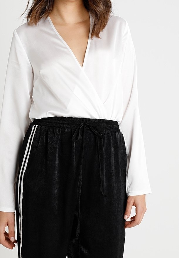 Femme portant un haut blanc à manches longues de style cache-cœur, associé à un pantalon noir velouté à cordon de serrage avec des bandes blanches sur les côtés.