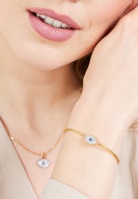 Gouden armband met een ovale, blauwe centrale versiering, omringd door kleine heldere stenen; gedragen om een pols tegen een lichtgekleurde stof.