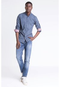 Blå knytblus med geometriskt mönster, uppvikta rutiga ärmar, tillsammans med ljusblåjeans och vita sneakers.