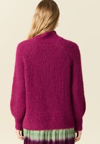 Fuksia neulepaita, jossa on korkea kaulus, väljä istuvuus ja ribbikuvio. Varusteena pitkät, hieman puffimaiset hihansuut.