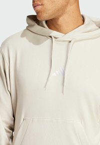 Hoodie beige chiaro, tessuto morbido, con tasca a canguro, cappuccio regolabile con cordoncino e logo ricamato in bianco sul petto.