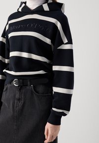 Sudadera negra con rayas blancas y logo bordado, combinada con una falda vaquera oscura y un cinturón negro con hebilla plateada.