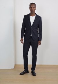 Un costume bleu marine foncé avec une veste sur mesure et un pantalon slim, associé à un t-shirt blanc et des chaussures en cuir noires sur un parquet en bois clair.