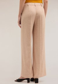 Szerokie, jasnopomarańczowe spodnie z pionowymi paseczkami, wykonane z teksturowanej tkaniny. Posiadają dopasowaną talię oraz subtelne detale szycia, połączone z czarnymi szpilkami.