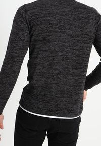 Pull gris foncé texturé avec un design ajusté et des manches longues, présentant un col rond et un effet superposé avec un ourlet blanc.