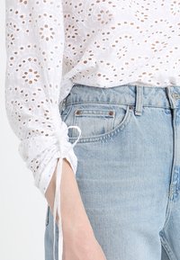 Hvid bluse med hulbroderi, runde mønstre og snøret ved manchetten, parret med lyseblå denim jeans.