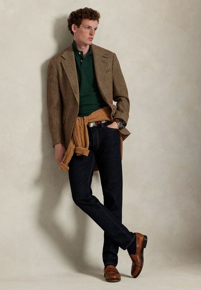 Jeune homme aux cheveux bouclés appuyé contre un mur, portant un blazer marron, un polo vert, un jean foncé, un pull noué et des chaussures en cuir marron.