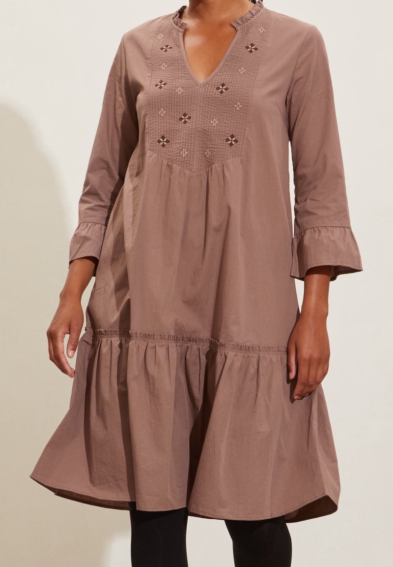 Femme portant une robe mauve ample, arrivant aux genoux, avec des manches 3/4, un ourlet froncé, un devant brodé et des leggings noirs en dessous.