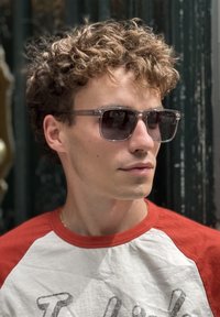 Ochelari de soare cu rame transparente și lentile întunecate. FațăCirculară, păr scurt și creț, purtând un tricou raglan roșu și alb. Detalii minime de fundal.