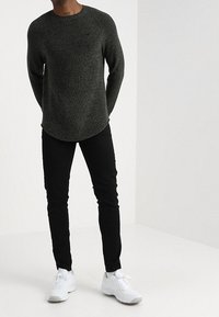 Mörkgrön stickad tröja med rundad nederkant, ihop med svarta slim-fit byxor och vita sneakers. Texturen verkar tjock och mysig.