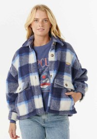 Blonde vrouw met een blauw-wit geblokt wollen jack over een blauwe graphic sweatshirt en lichtblauwe jeans, staand met één hand in de zak.