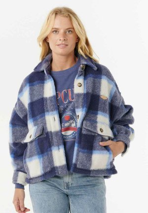 Blonde vrouw met een blauw-wit geblokt wollen jack over een blauwe graphic sweatshirt en lichtblauwe jeans, staand met één hand in de zak.