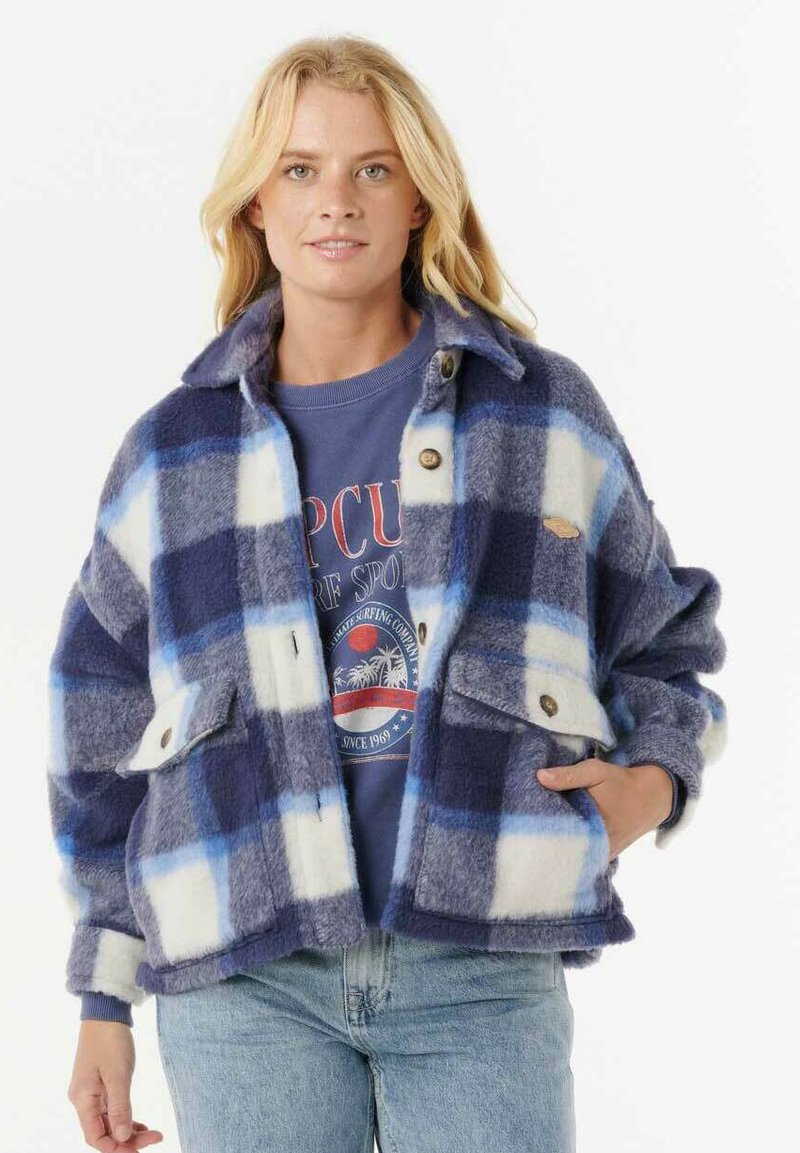 Blonde vrouw met een blauw-wit geblokt wollen jack over een blauwe graphic sweatshirt en lichtblauwe jeans, staand met één hand in de zak.