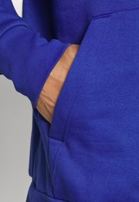 Main placée dans la poche avant d'un sweat à capuche bleu vif avec une texture de tissu lisse.