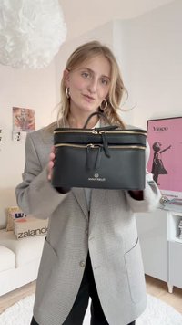 Zwarte leren tas met gouden ritsen, rechthoekige vorm, dubbele compartimenten en een handvat aan de bovenkant. Voorzien van een logo aan de voorkant.