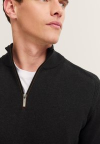Maglione zip-up nero con colletto alto, realizzato in tessuto a maglia testurizzato. Presenta una cerniera argentata e un discreto logo ricamato sul petto.
