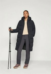 Άτομο στέκεται κρατώντας μαύρους μπαστούνες πεζοπορίας, φορώντας ένα μακρύ μαύρο μπουφάν, μπεζ μπλούζα, σκούρους παντελόνες και μωβ μποτάκια πεζοπορίας σε μια απλή φόντο.