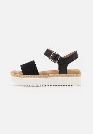 Sort platform sandal med bred frontstrop, ankelstrop med gyldent spænde, flettet mellemsål og tyk hvid rillede sål.