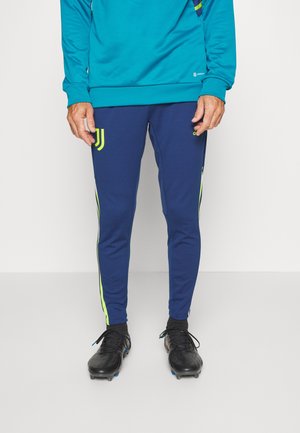 Homme portant un pantalon de sport bleu avec des bandes néon, des chaussures à crampons noires et un haut turquoise, debout sur un fond blanc uni.