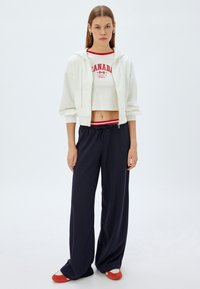 Hvid cropped hoodie med lynlås foran, parret med en rød og hvid Canada-logo top, marineblå vide bukser og røde sportsko.