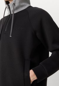 Sudadera negra con cremallera y capucha gris, con una tela texturizada, bolsillo delantero y logo bordado en el pecho.