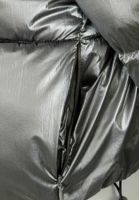 Bundička s kapucňou v metalickej šedej tkanine s hladkou textúrou. Obsahuje zipsovú kapsu, prešívanie a jemný lesk.