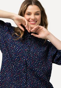 Námořnická košile na knoflíky s vzorem malých barevných tvarů v červené, béžové a modré; má klasický límec a krátké rukávy.