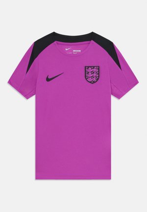 Violeta sporta krekls ar melniem akcentiem, uz kura ir melna Nike logo un emblēma uz krūšu daļas. Īsas piedurknes un teksturēta auduma.