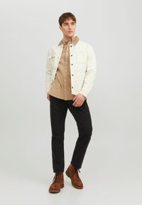 Chaqueta de mezclilla blanca sobre una camisa de botones beige, pantalones negros y botas marrones. La chaqueta tiene bolsillos delanteros y botones negros.